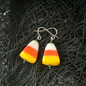 Pode incluir: Um par de brincos pendentes com contas de vidro em forma de milho doce com listras amarelas, laranja e brancas. Os brincos têm ganchos prateados.