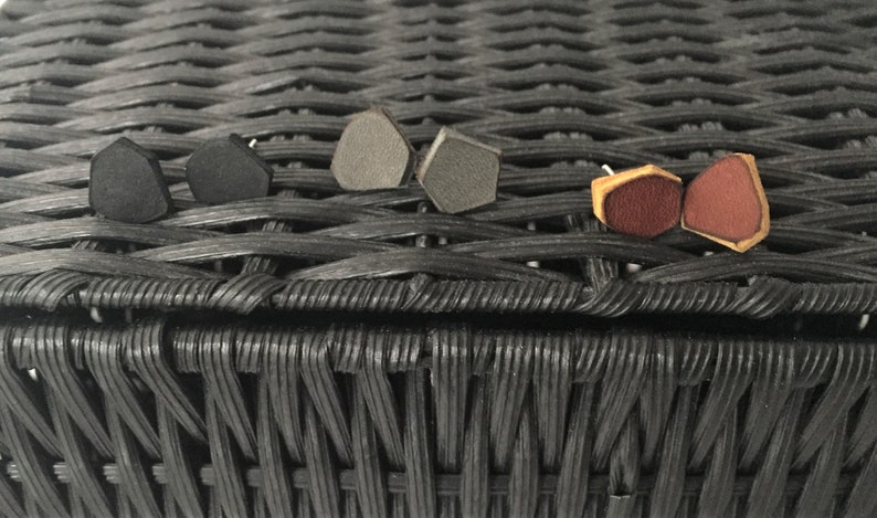 May include: Assortment of geometric shaped earrings in black, gray, and brown leather. The earrings are displayed on a black wicker surface.