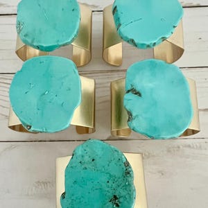 May include: A set of five napkin rings, each featuring a large, irregular, polished turquoise-colored stone slab mounted onto a wide, smooth, matte gold-toned metal band. The stones display natural variations in shape, texture, and color.