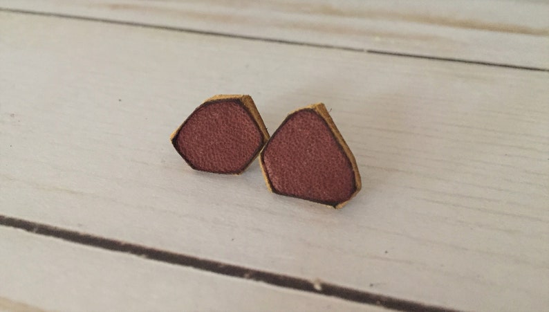 May include: A pair of small, hexagonal shaped earrings made of leather. The earrings are a deep burgundy color with a light brown border.