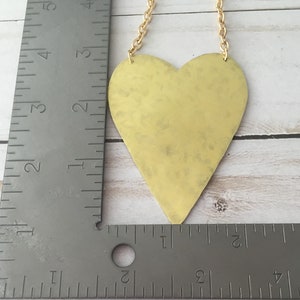 Op de afbeelding: Een gouden hartvormige hanger hangt aan een gouden ketting. De hanger is ongeveer 7,5 cm hoog en 7,5 cm breed. De hanger heeft een gehamerde textuur.
