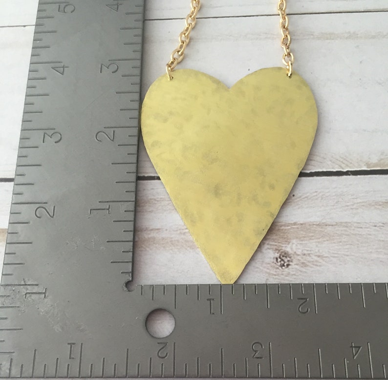 Op de afbeelding: Een gouden hartvormige hanger hangt aan een gouden ketting. De hanger is ongeveer 7,5 cm hoog en 7,5 cm breed. De hanger heeft een gehamerde textuur.