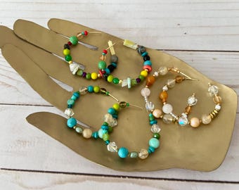 May include: Four pairs of beaded hoop earrings in various colors, including turquoise, green, yellow, and pink. The earrings are displayed on a gold-colored hand-shaped tray against a light wood background.