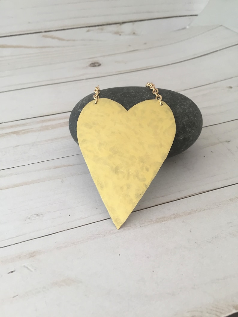 Op de afbeelding: Een grote, goudkleurige hartvormige ketting met een gehammerde textuur. De hanger hangt aan een gouden ketting.