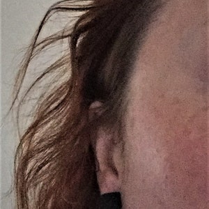May include: Close-up of a woman's ear with a small, dark earring. Her hair is long and brown, and her skin is fair.