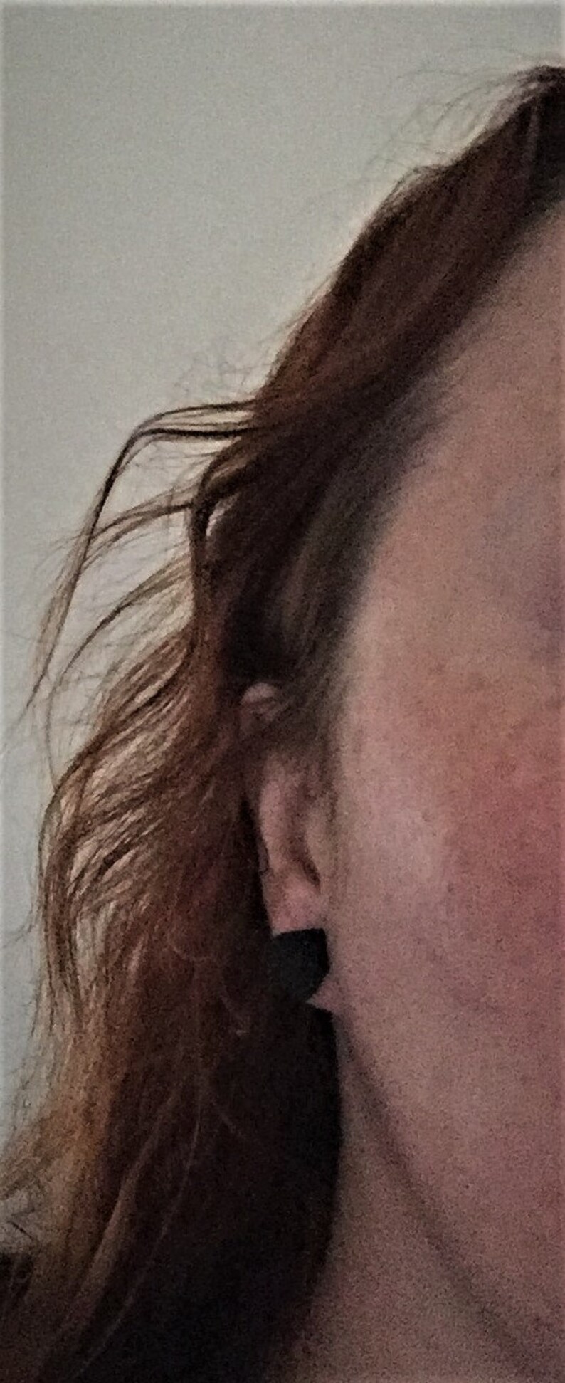 May include: Close-up of a woman's ear with a small, dark earring. Her hair is long and brown, and her skin is fair.