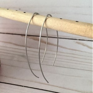 Puede incluir: Un par de pendientes de aro de plata con un diseño simple y curvo. Los pendientes se muestran colgando de una varilla de madera.