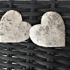 May include: Two white heart-shaped metal ornaments with a distressed finish, resting on a black woven wicker surface.