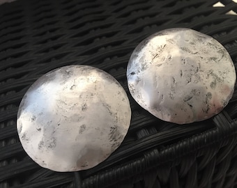 May include: Two silver-toned metal discs with a textured surface. The discs are round and have a slightly raised center.
