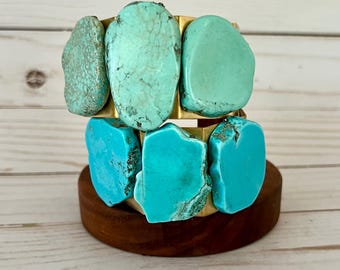 May include: A gold-toned metal cuff bracelet featuring three large, irregular, polished turquoise stones in shades of light blue and green. The stones are set horizontally across the front of the band, showcasing natural veining and smooth, rounded edges.