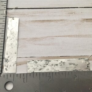 Puede incluir: Un par de pendientes de plata con forma rectangular y textura martillada. Los pendientes cuelgan de una regla.
