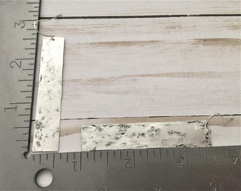 Puede incluir: Un par de pendientes de plata con forma rectangular y textura martillada. Los pendientes cuelgan de una regla.