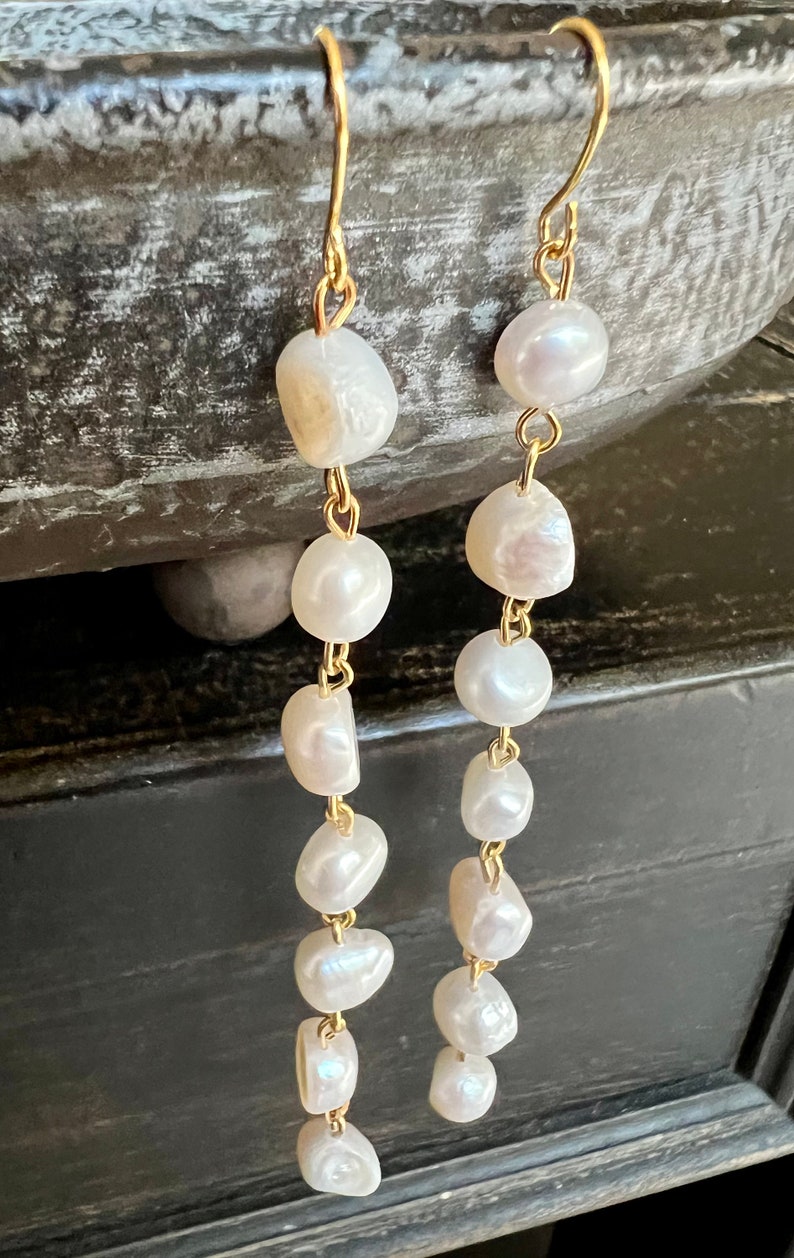 Pu&ograve; includere: Un paio di lunghi orecchini pendenti dorati con perle bianche. Gli orecchini hanno un design semplice con una catena e pi&ugrave; perle.
