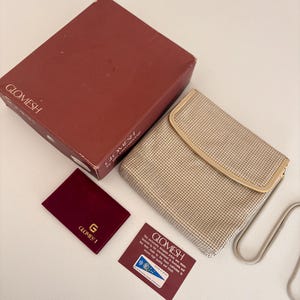 Puede incluir: Un bolso Glomesh vintage con dise&ntilde;o de malla, una caja a juego y una peque&ntilde;a bolsa burdeos. El bolso tiene una correa de cadena plateada y un marco beige. El logotipo de Glomesh est&aacute; presente.