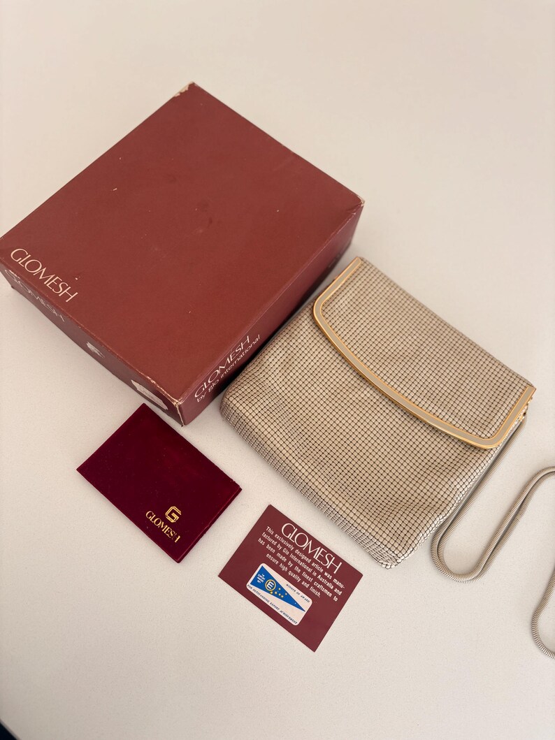 Puede incluir: Un bolso Glomesh vintage con dise&ntilde;o de malla, una caja a juego y una peque&ntilde;a bolsa burdeos. El bolso tiene una correa de cadena plateada y un marco beige. El logotipo de Glomesh est&aacute; presente.