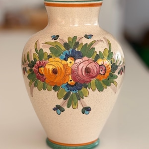 May include: Cream-coloured ceramic vase with a floral design. The vase features a bouquet of colourful flowers in shades of orange, pink, blue, and yellow, with green leaves. The rim and base are trimmed with green and orange bands.