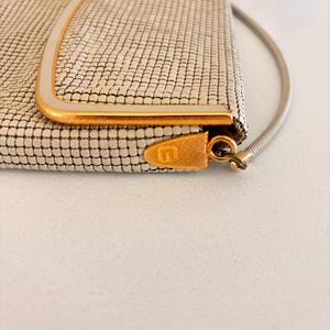 Puede incluir: Un bolso de mano vintage de malla blanquecina con marco y cierre dorados. El bolso rectangular presenta un delicado patr&oacute;n tejido y una fina correa de cadena plateada. El cierre est&aacute; grabado con una letra estilizada "U".