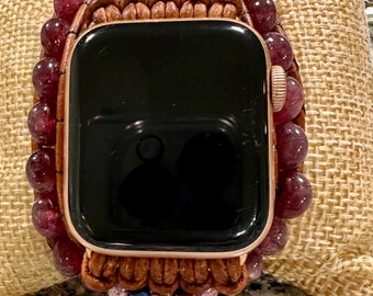 May include: An Apple Watch with a rose gold case and a custom beaded band. The band features dark red, round beads, brown leather accents, and small, colorful crystal embellishments. The watch is displayed on a tan, textured surface.