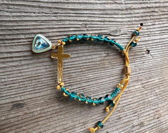 May include: A gold-colored cross charm bracelet with teal and gold accents. The bracelet features a cross pendant and a triangular charm with a blue stone. The bracelet is made with teal beads and gold-colored cord.
