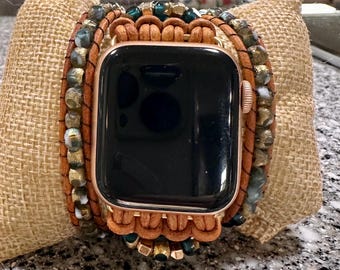 May include: A rose gold Apple Watch with a brown leather wrap bracelet. The bracelet is adorned with various beads in shades of green, gold, and white. The watch is resting on a burlap pillow.