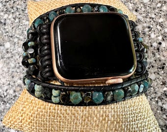 May include: A black leather wrap watch band with teal and black beads. The band is wrapped around a gold Apple Watch. The watch and band are resting on a small tan burlap pillow.
