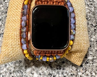 May include: A rose gold smartwatch with a black screen is displayed on a burlap pillow. The watch is adorned with a wrap-around band featuring brown leather and beaded accents in yellow, purple, and gold.