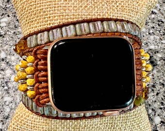 May include: A brown leather wrap watch band with a rose gold watch face. The band features yellow and gray beads, and a woven design. The watch is displayed on a tan burlap pillow.