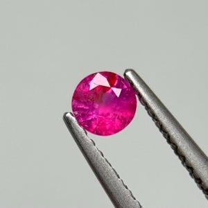 Könnte beinhalten: Ein runder, facettierter Rubin-Edelstein, der von einer silbernen Pinzette gehalten wird. Der Rubin hat eine leuchtende, rosarote Farbe mit sichtbaren inneren Einschlüssen. Die Pinzette ist fokussiert.