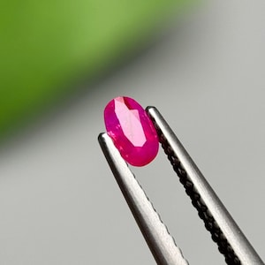 Puede incluir: Una gema de rubí facetada, de forma ovalada, sujeta con unas pinzas plateadas. El rubí es de un color rojo-rosa intenso y vibrante, con múltiples facetas que captan la luz. El fondo es un degradado suave de verde y gris.