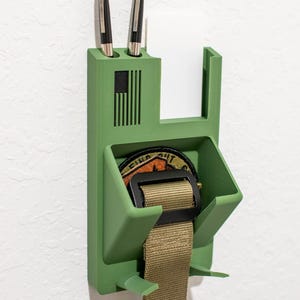May include: A green wall-mounted organizer with two pens, a notepad, and a compartment for a belt and patch. The organizer features an American flag design and is made of a solid, matte material.