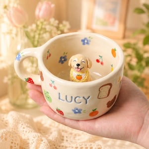 May include: Cream-colored ceramic mug with a smiling puppy figurine inside. The mug is decorated with colorful fruit and flower illustrations, and the name "LUCY" is written on the side. The handle is adorned with a strawberry and blue flowers.
