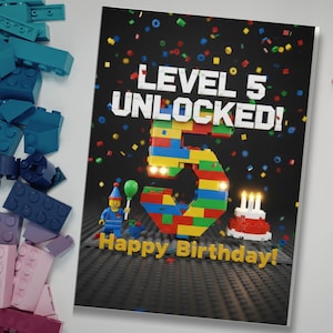 May include: A birthday card with the text "LEVEL 5 UNLOCKED!" and "Happy Birthday!". The number 5 is constructed from colorful toy blocks. A toy figure holds a green balloon. A small cake with candles is also present.