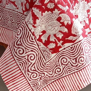 Peut inclure: Une nappe à motifs floraux rouges et blancs. Le motif présente de grands motifs floraux blancs et des motifs tourbillonnants sur un fond rouge. La bordure présente un motif à rayures. La nappe est drapée sur une table.