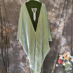 May include: A sage green velvet kimono-style garment featuring a deep V-shaped back panel in a darker forest green velvet. The back panel is embellished with a triangular beaded patch featuring dangling white fringe accents at the center.
