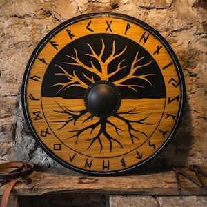 Pode incluir: Escudo redondo de madeira com um desenho de árvore da vida em preto e amarelo. O escudo tem uma borda preta e um chefe central preto. O design inclui símbolos rúnicos. Uma correia de couro é visível.
