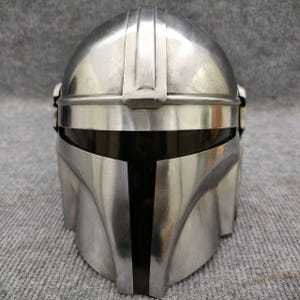 Puede incluir: Un casco plateado de Mandaloriano con una visera negra. El casco tiene una superficie lisa y reflectante con crestas verticales en la parte superior. El dise&ntilde;o recuerda a un personaje espacial.