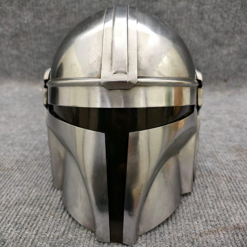 Puede incluir: Un casco plateado de Mandaloriano con una visera negra. El casco tiene una superficie lisa y reflectante con crestas verticales en la parte superior. El dise&ntilde;o recuerda a un personaje espacial.