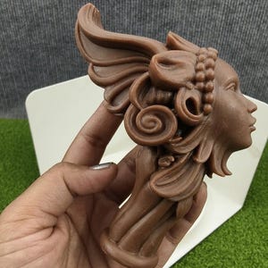May include: A brown bust of a woman with ornate hair and headwear. The sculpture features detailed curls, a bow, and a beaded band. The bust is held in a hand against a white and green background.
