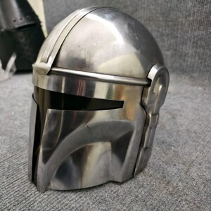 Puede incluir: Un casco plateado con visera, con parte superior redondeada y placa frontal angular. El casco tiene una superficie lisa y reflectante y detalles laterales, lo que sugiere un tema futurista o de ciencia ficci&oacute;n.