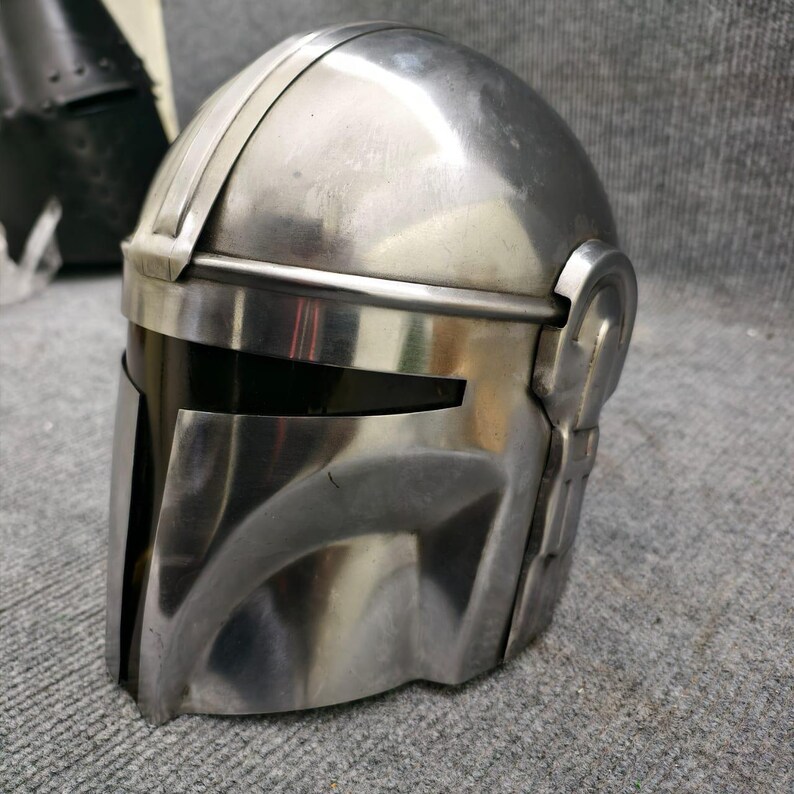 Puede incluir: Un casco plateado con visera, con parte superior redondeada y placa frontal angular. El casco tiene una superficie lisa y reflectante y detalles laterales, lo que sugiere un tema futurista o de ciencia ficci&oacute;n.