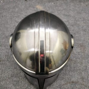 Puede incluir: Un casco plateado con una parte superior redondeada y crestas verticales. El casco tiene una superficie reflectante y un &aacute;rea de visera. El dise&ntilde;o sugiere un tema futurista o de ciencia ficci&oacute;n, posiblemente para cosplay o exhibici&oacute;n.