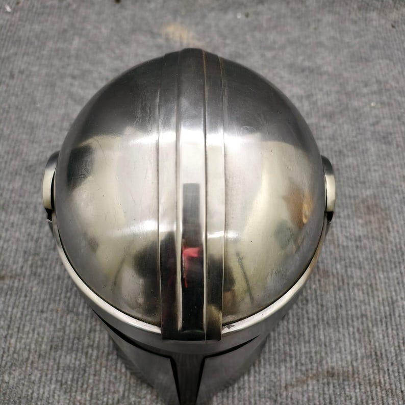 Puede incluir: Un casco plateado con una parte superior redondeada y crestas verticales. El casco tiene una superficie reflectante y un &aacute;rea de visera. El dise&ntilde;o sugiere un tema futurista o de ciencia ficci&oacute;n, posiblemente para cosplay o exhibici&oacute;n.
