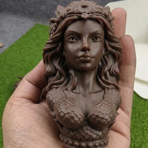 May include: A brown bust of a woman with long wavy hair, a crown, and a scaled bodice. The sculpture is held in a hand, with a green and white background.