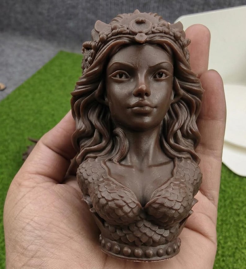 May include: A brown sculpture of a woman's bust, featuring long wavy hair, a crown, and a scaled bodice. The bust is held in a hand, with a green and white background.