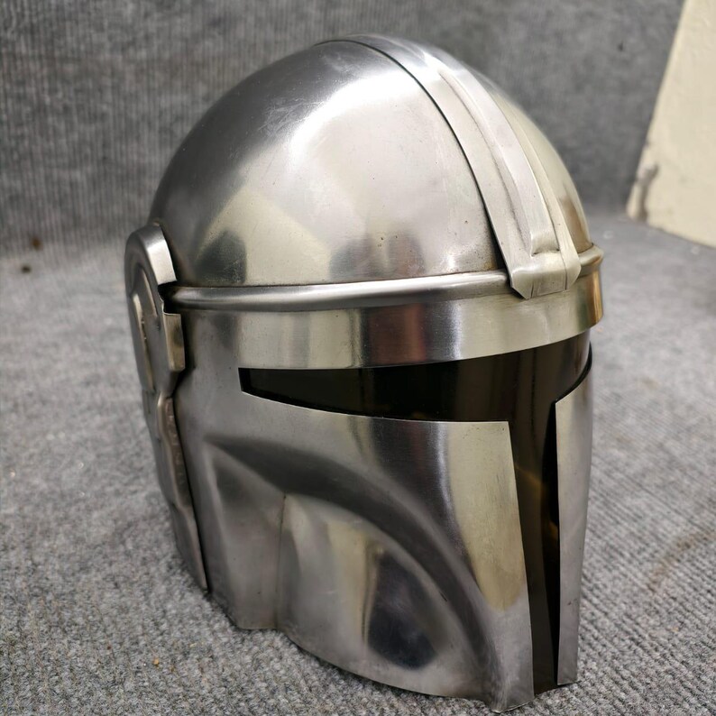 Puede incluir: Un casco plateado met&aacute;lico con visera, con una distintiva abertura de visera en forma de T. El casco tiene una superficie lisa y reflectante y una prominente cresta que recorre la parte superior. El dise&ntilde;o sugiere un tema futurista o de ciencia ficci&oacute;n.