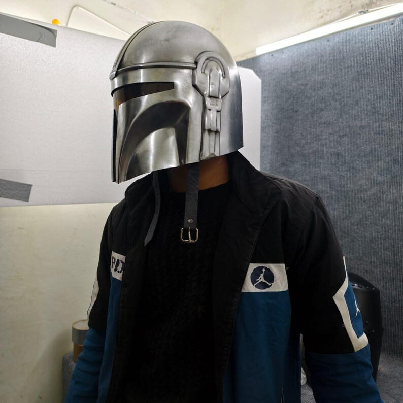 Puede incluir: Un casco plateado de Mandaloriano con visera y carrilleras. El casco tiene una correa y se lleva sobre una chaqueta negra con detalles azules y un logotipo.