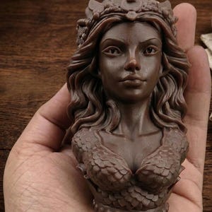 May include: A brown sculpture of a woman's bust, featuring a crown, flowing hair, and a scaled top. The detailed artwork is held in a hand. The bust is a decorative item, with intricate details.