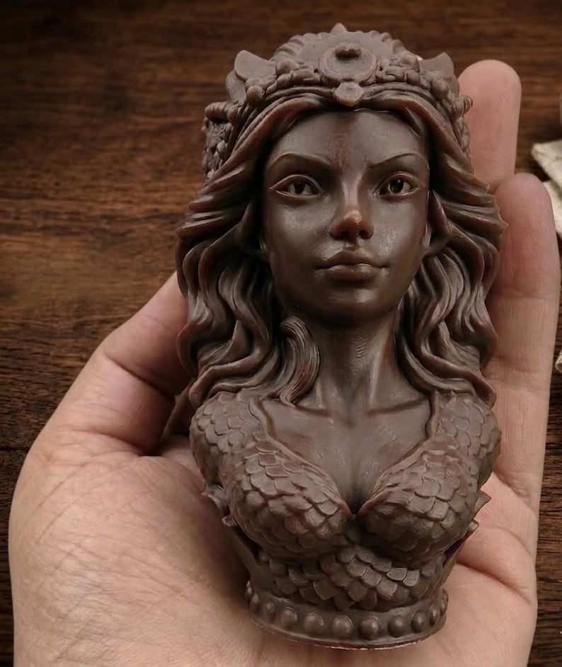 May include: A brown sculpture of a woman's bust, featuring a crown, flowing hair, and a scaled top. The detailed artwork is held in a hand. The bust is a decorative item, with intricate details.