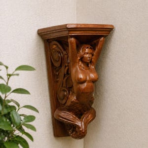 Puede incluir: Estante de esquina de madera con una figura de sirena tallada. La sirena sostiene el estante con los brazos levantados. El estante es de color marrón cálido y está montado en una esquina contra una pared neutra.