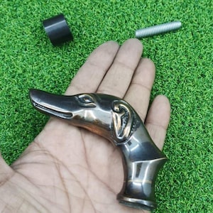 May include: A bronze-toned dog head cane handle. The handle is detailed with the dog's features, including the eyes, ears, and snout. A black cylinder and a silver screw are also visible. The handle is held in a hand against a green background.